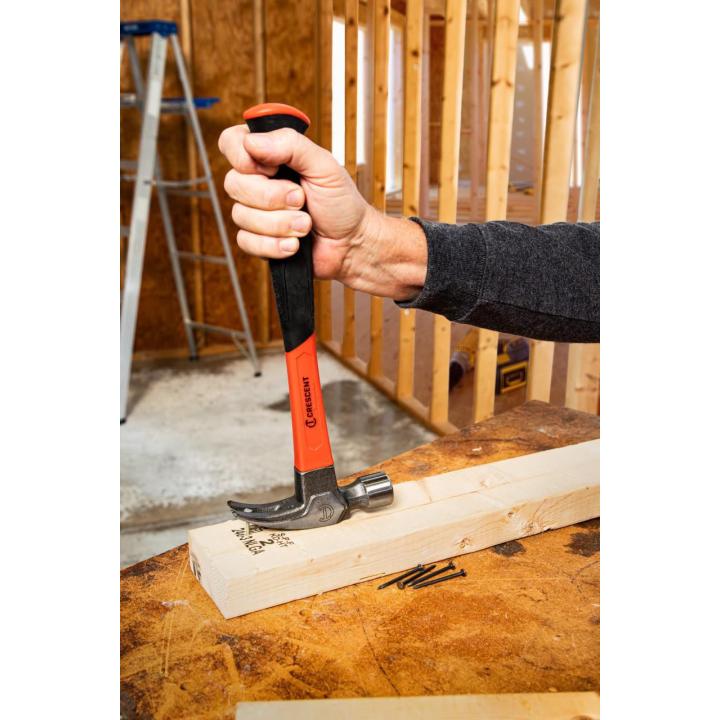 Worker grips a Crescent hammer vertically over a board, with its head beside loose nails in unfinished framing.