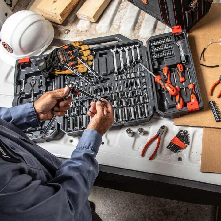 180 Piece Mechanics Tool Set in use on a workbench, with the open carrying case displaying organized sockets, wrenches, ratchets, screwdrivers, pliers, bits, and hex keys.