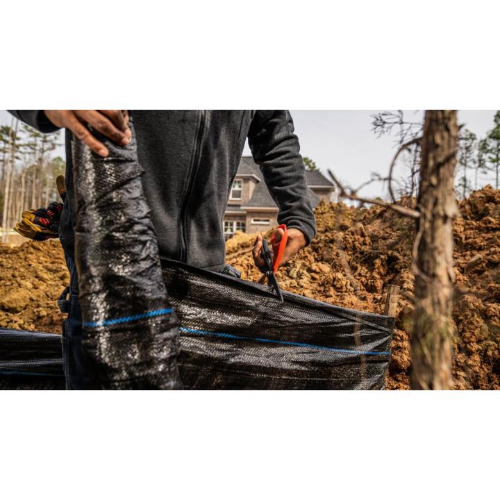 Person holding shears cutting a plastic tarp outdoors, with a house, soil and trees in the background.