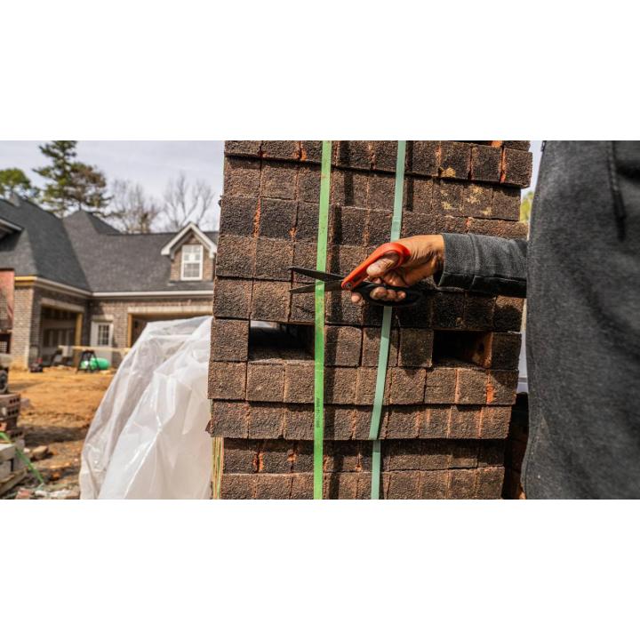 Hand holding orange-handled shears cutting green straps around stacked brown bricks at a construction site.