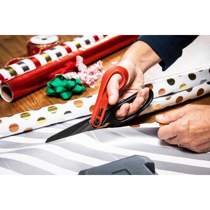 Hands cutting silver striped wrapping paper with red and black shears, with holiday wrapping paper rolls and green and red bows nearby.