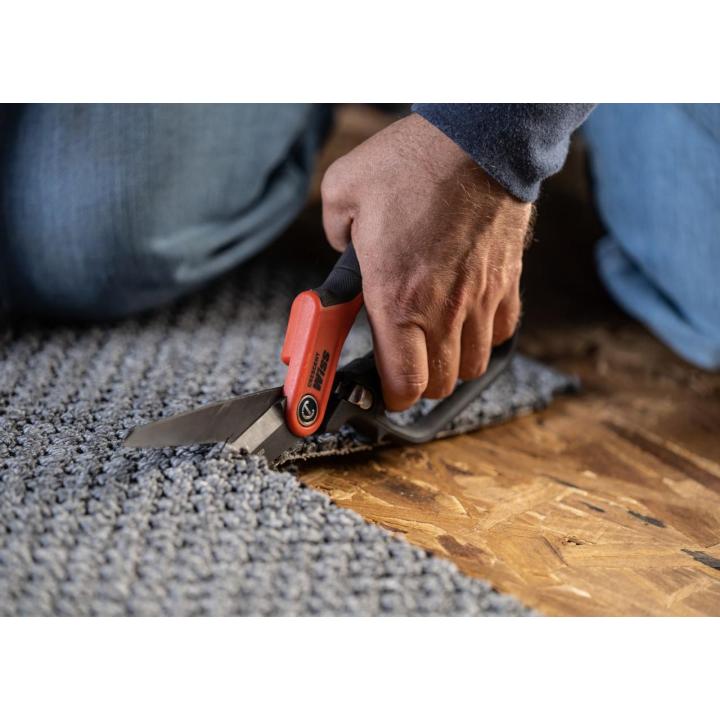 Person kneeling and cutting textured gray carpet with utility shears, revealing a wooden floor underneath.