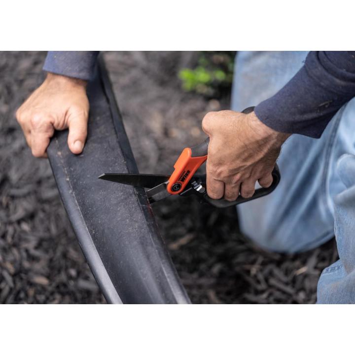 Hands cutting a black flexible irrigation tube with orange-handled shears over mulch-covered ground.