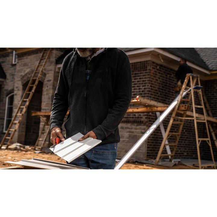 Worker cuts white colored soffit with shears at a construction site with wooden ladders and brick walls in the background.