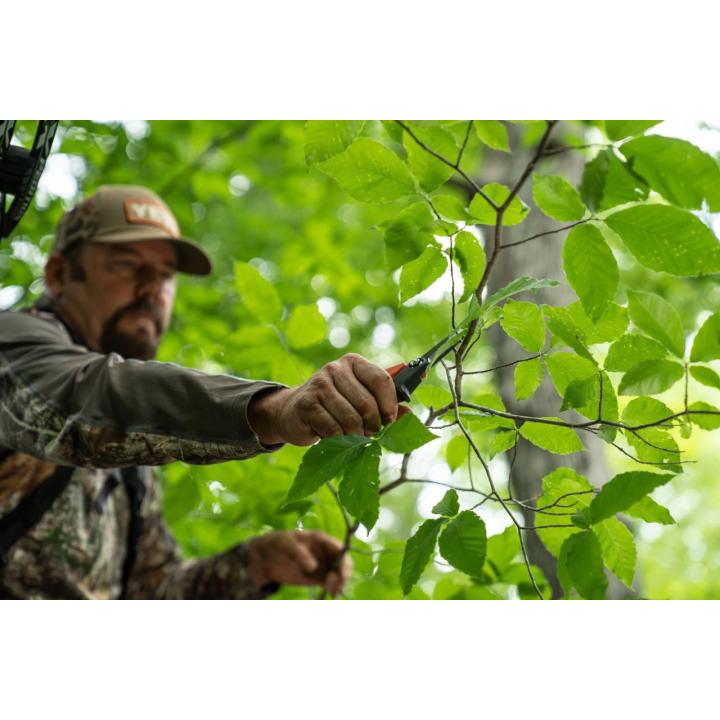 Man in a camouflage jacket and cap pruning a small tree branch with shears in a leafy green forest.