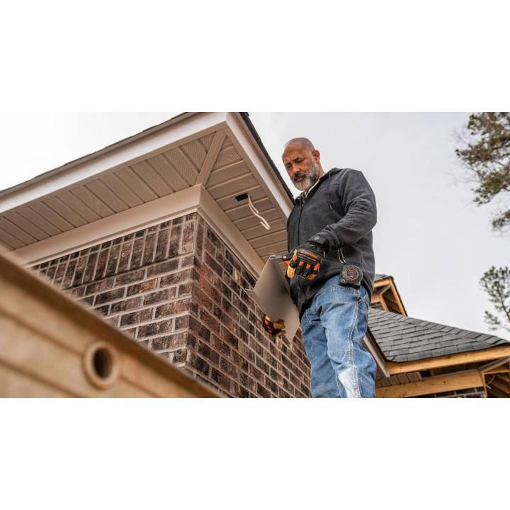 Man in work gloves cutting roofing material near the corner of a brick and wood house exterior under an overcast sky.