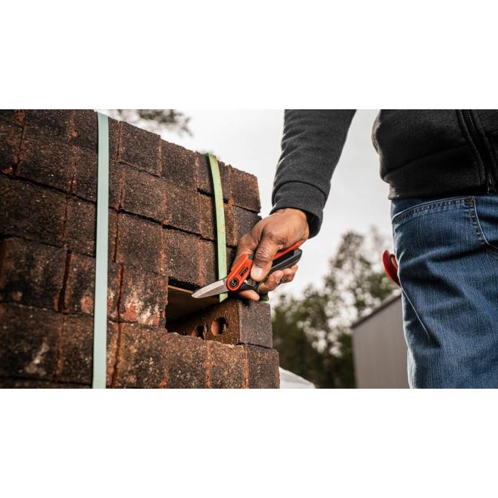 Person using a Crescent shear to slice a green strap securing a stack of brown bricks outdoors.